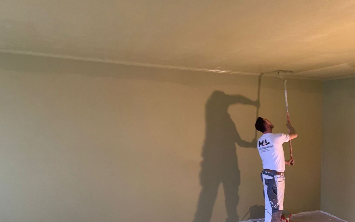 MEL-Maler streicht mit der Rolle eine Decke über einer salbeifarbenen Wand